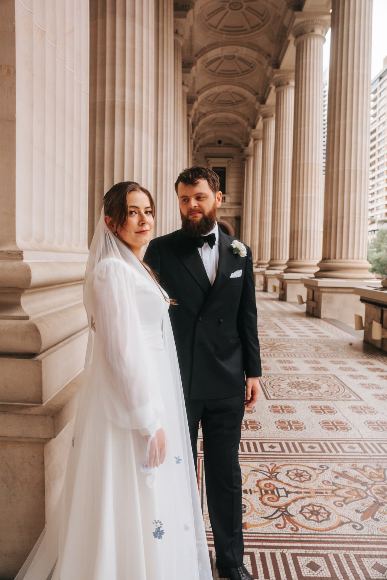 Bianca and Aidan standing beside a column in the colonnade of Parliament House, patterned tiles at their feet