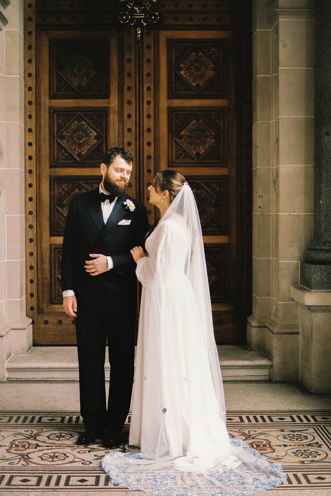 Bianca and Aidan at the carved timber doors of Parliament House, her veil and train over the tiles, on 35mm film