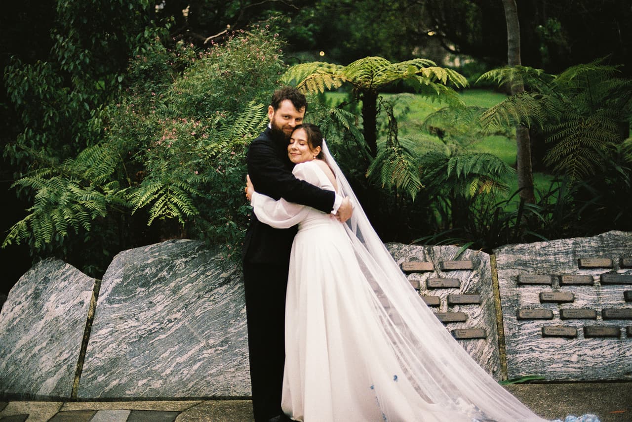 Bianca and Aidan embracing among tree ferns, her veil falling around them, on 35mm film