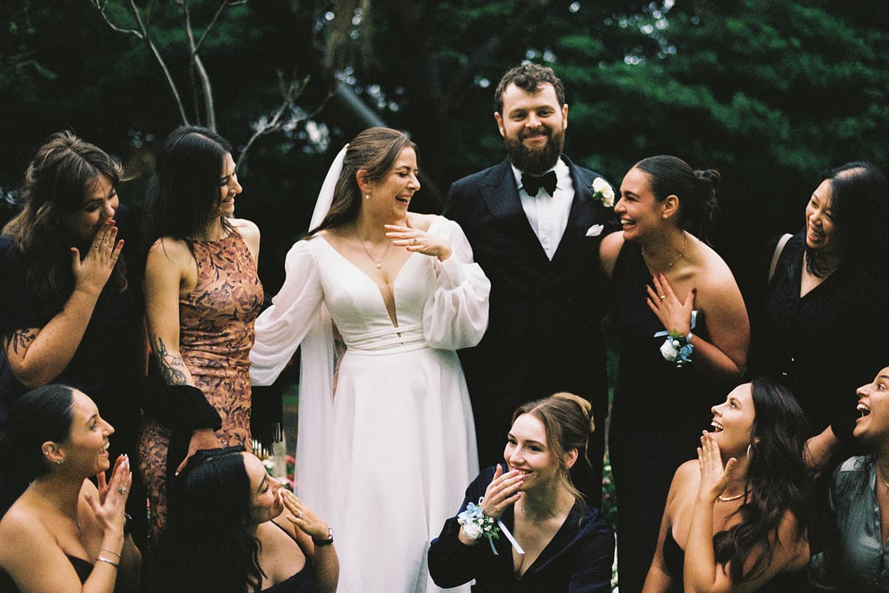 Bianca and Aidan laughing with a group of friends in dark dresses in the gardens, on 35mm film