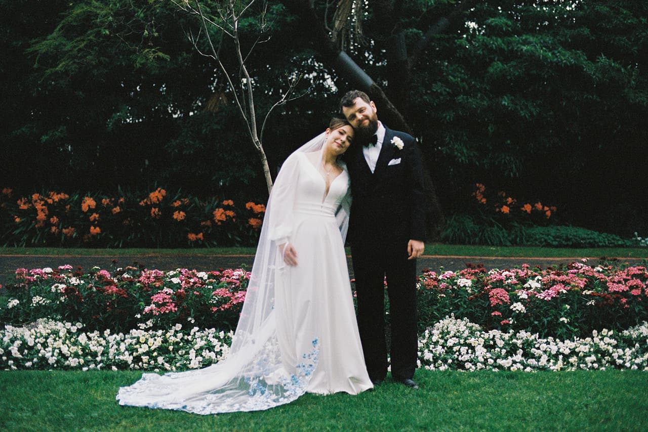 Bianca's long train spread over the grass as she and Aidan stand beside a flower bed, on 35mm film
