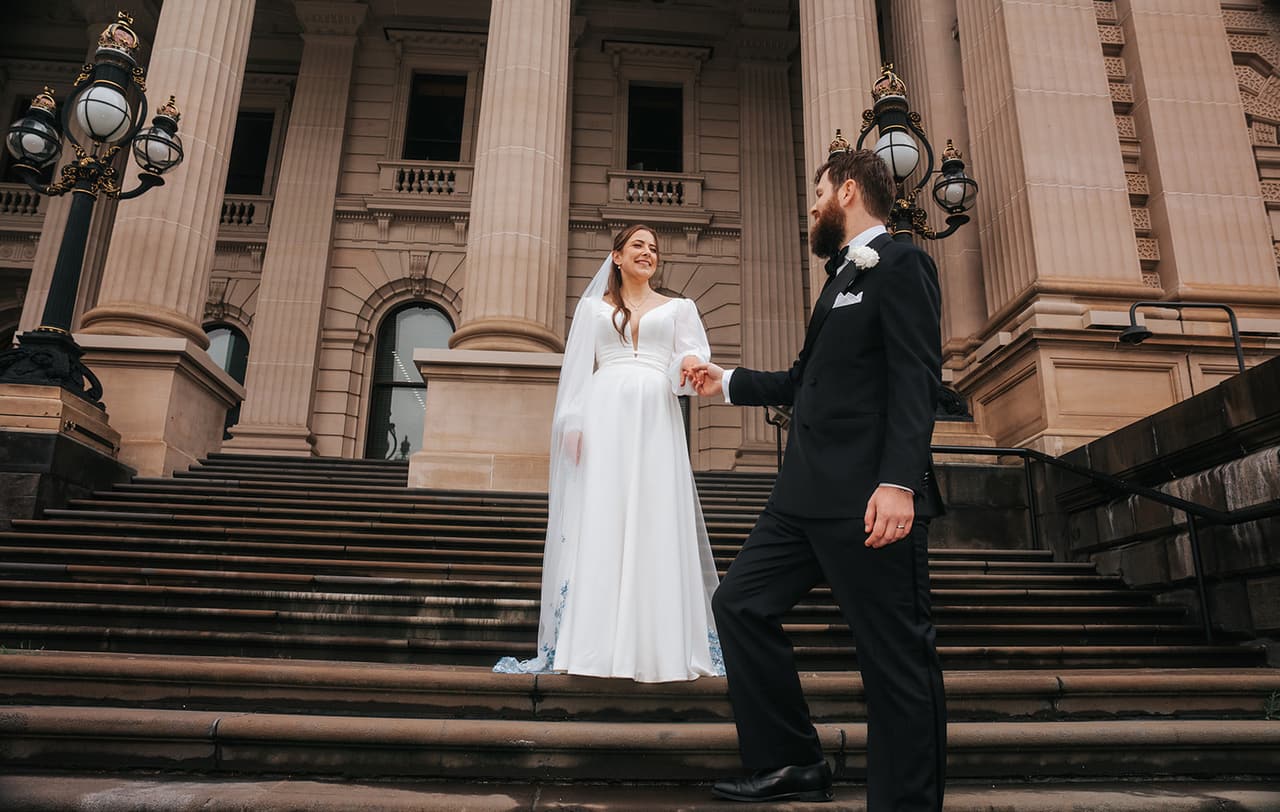Bianca and Aidan holding hands on the wide steps of Parliament House between the old lamps