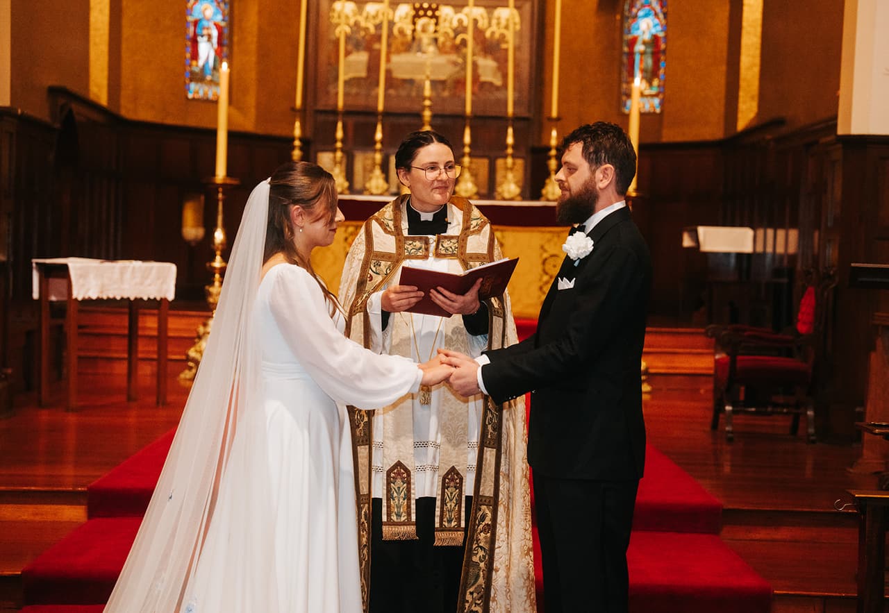 The priest reading as Bianca and Aidan hold hands before the altar, red carpet under their feet