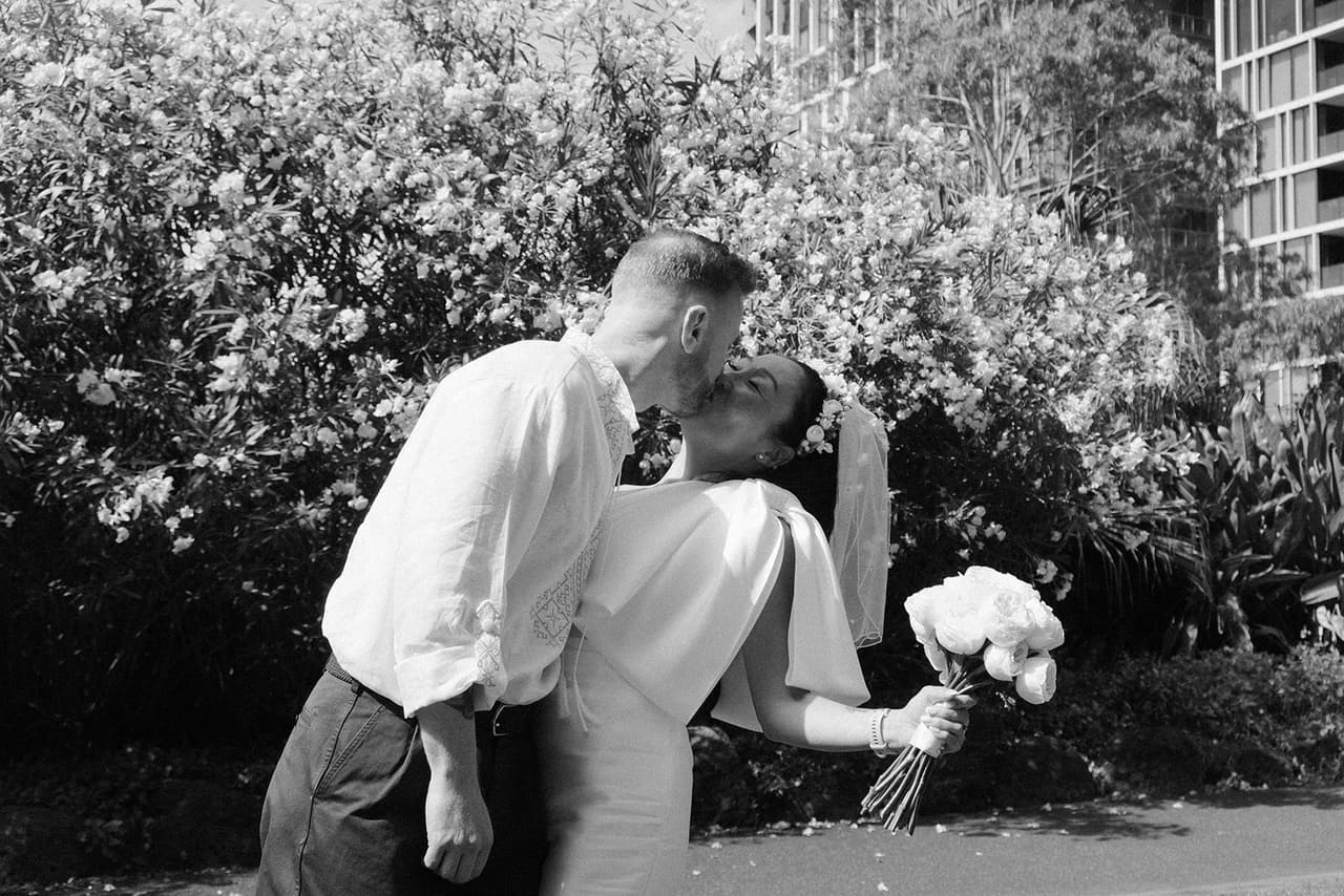 Black and white photo of Stefan leaning in to kiss Cecile beside a flowering hedge