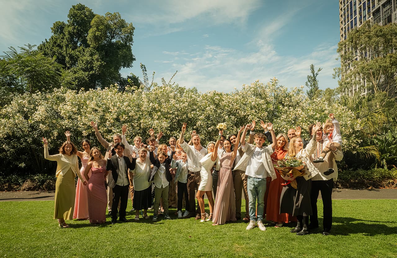 Cecile, Stefan and all their guests cheering with their arms in the air on the lawn in front of the flowering hedge