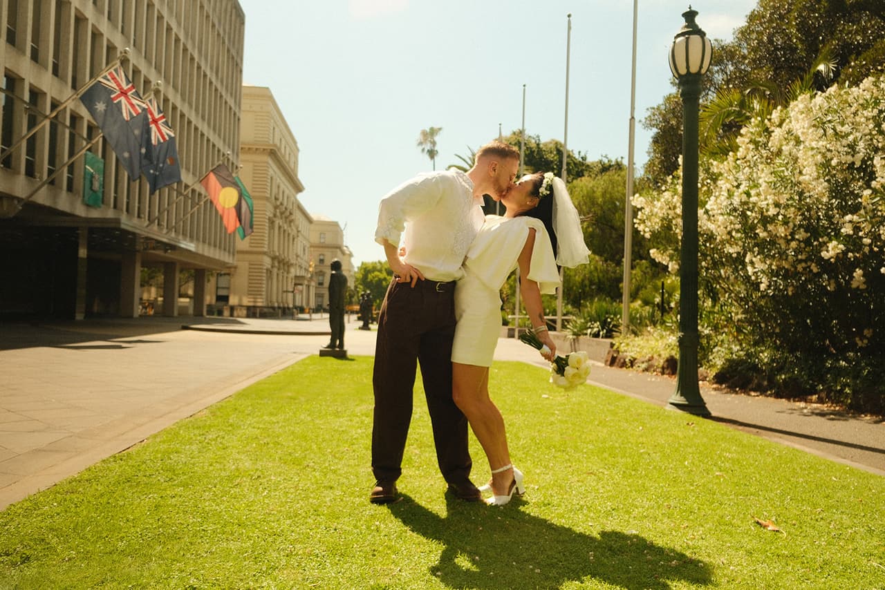 Cecile and Stefan kissing on a strip of lawn among the old government buildings, flags flying behind them