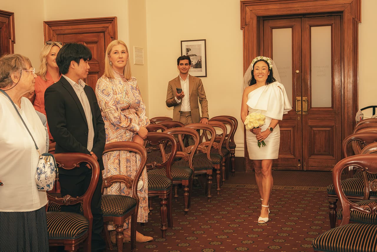 Cecile walking into the ceremony room past the rows of timber chairs as the guests stand and watch