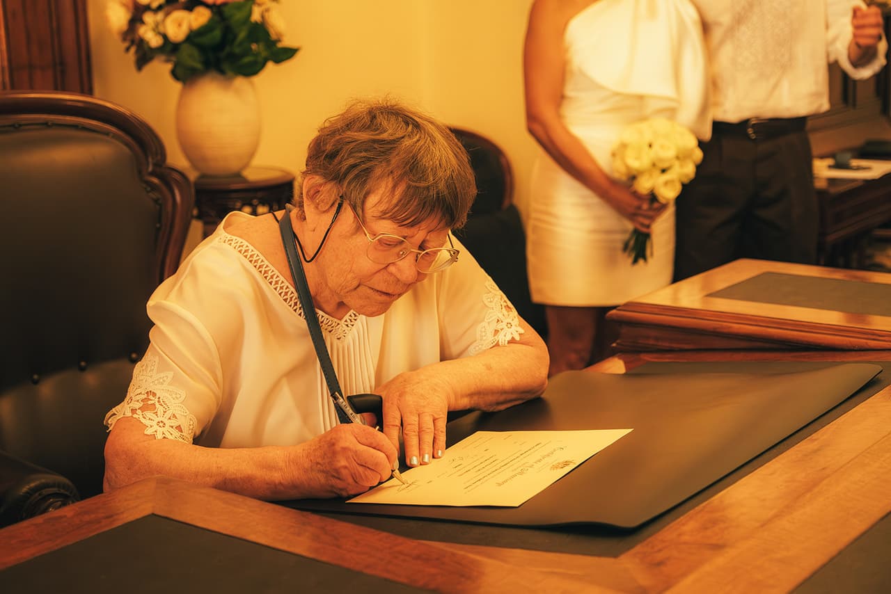 An older guest in a lace trimmed blouse signing at the registry desk while Cecile and Stefan stand by