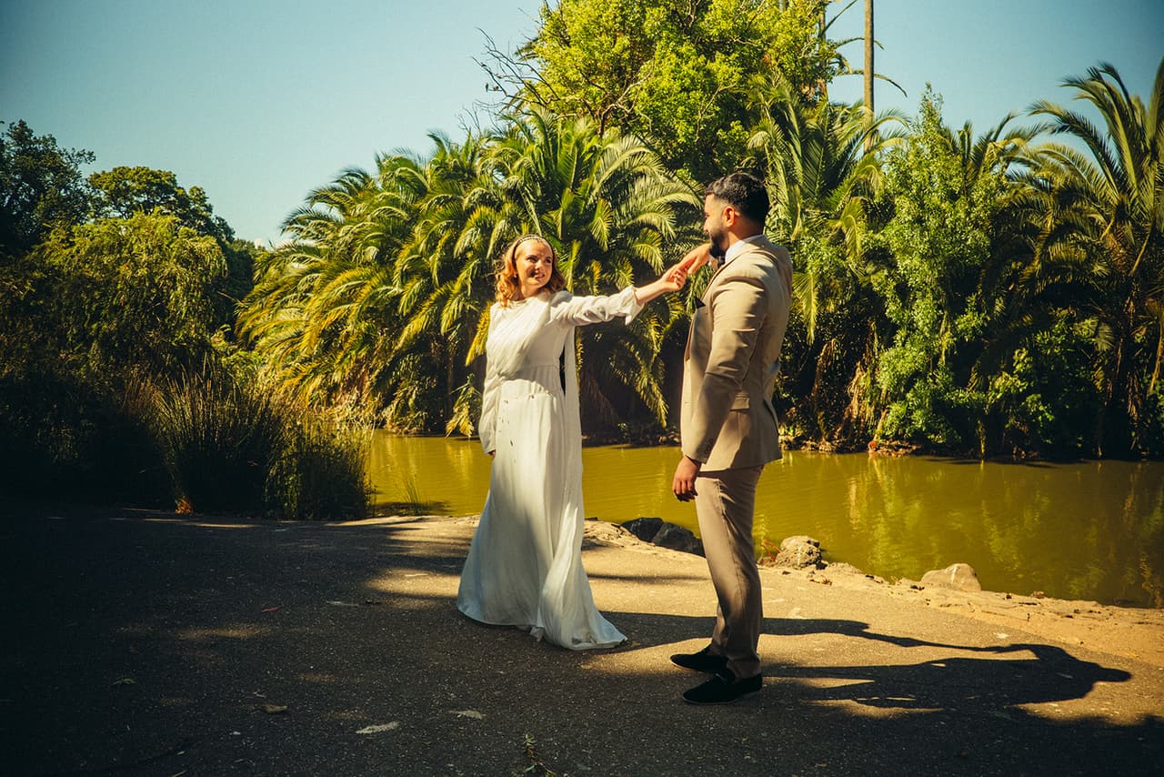 Elektra laughing and reaching for Yasir’s face as they dance on the path beside the water and the palms