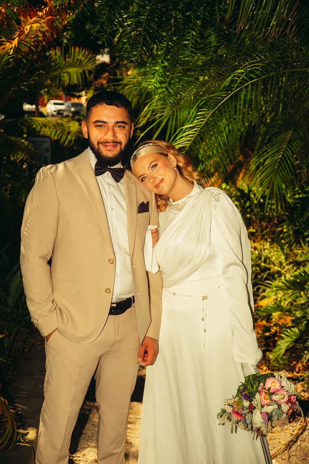 Elektra resting her head on Yasir’s shoulder on a path lined with ferns and palms