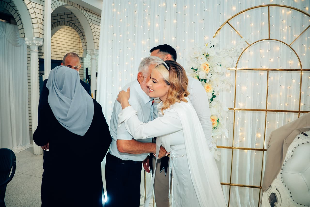 An older man hugging Elektra by the head table as an older woman in a grey headscarf looks on