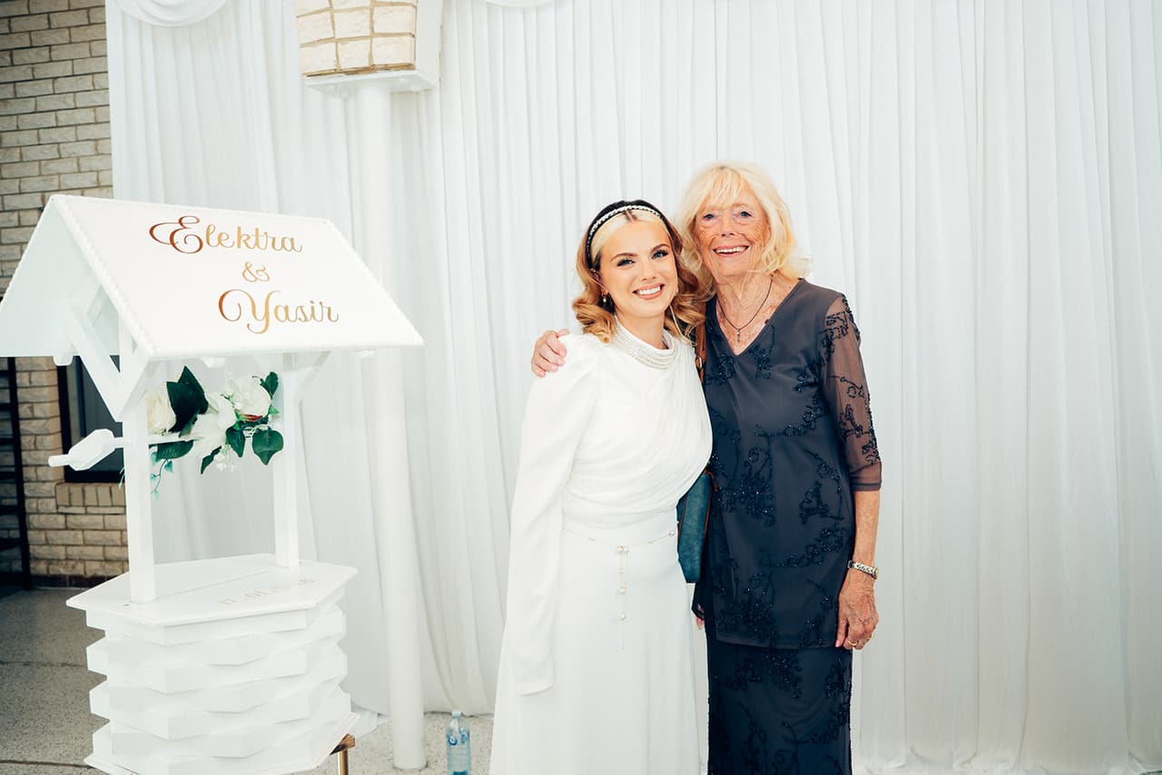 Elektra with her arm around an older woman in a dark lace dress beside the white wishing well