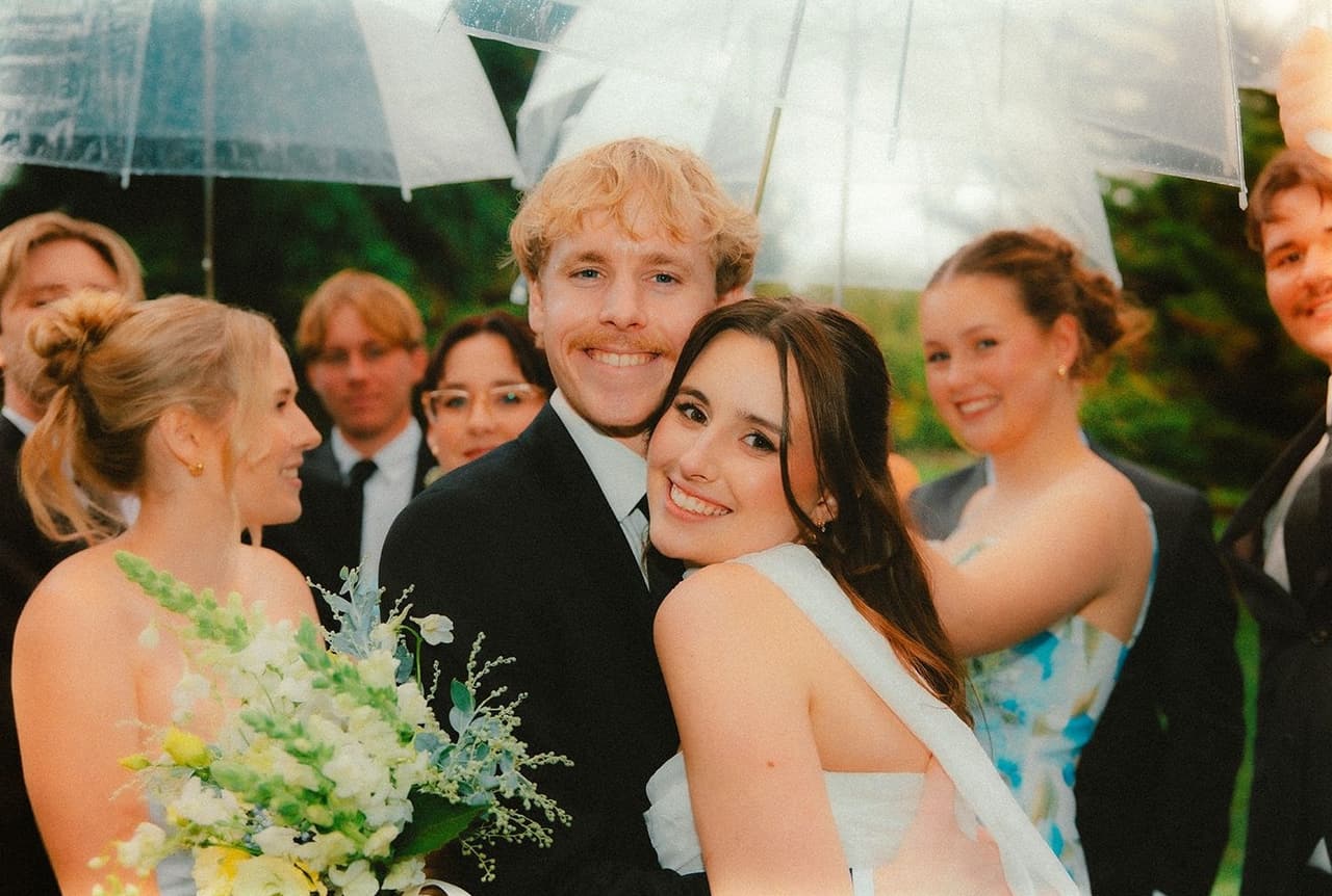 Eli and Charlotte laughing with friends under clear umbrellas after the ceremony