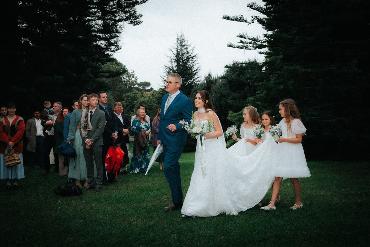 Charlotte walking in on her father’s arm past the standing guests, three flower girls carrying her train