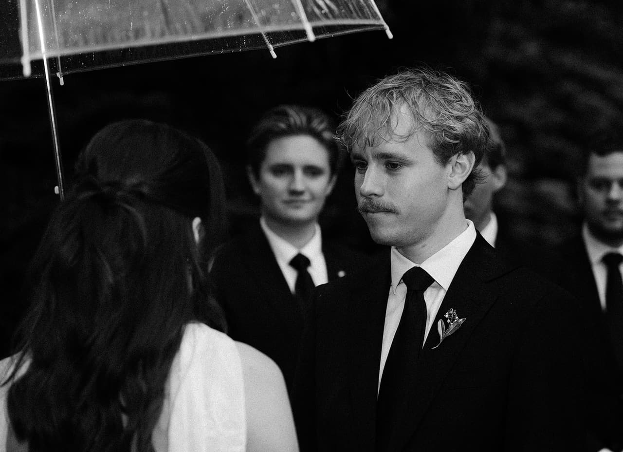 Black and white photo of Eli looking at Charlotte during the vows, a groomsman smiling behind him