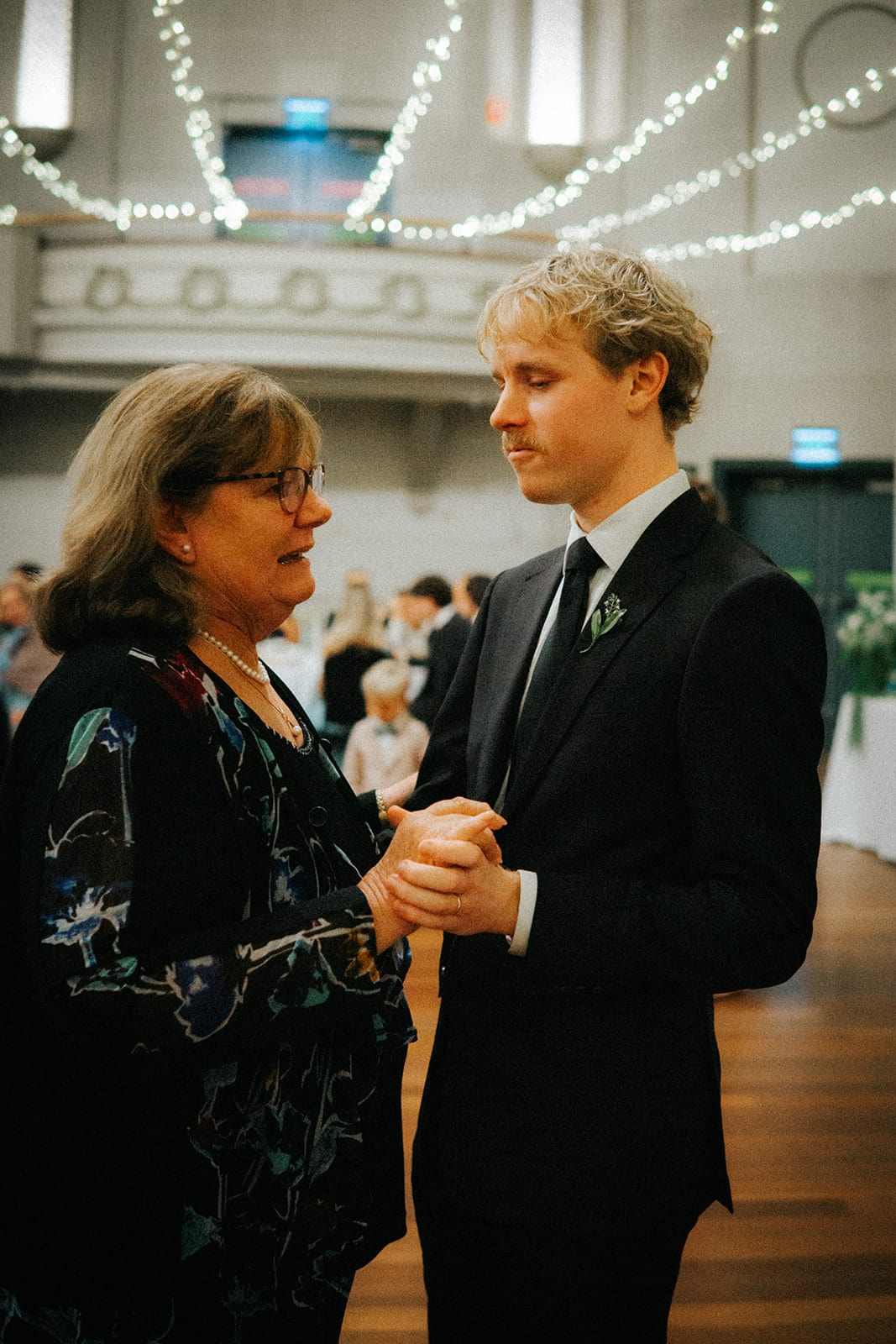 Eli dancing with a woman in a dark floral dress under the fairy lights of the hall