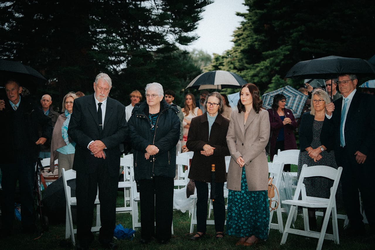 Family and friends standing among the white chairs to watch the ceremony, umbrellas up behind them