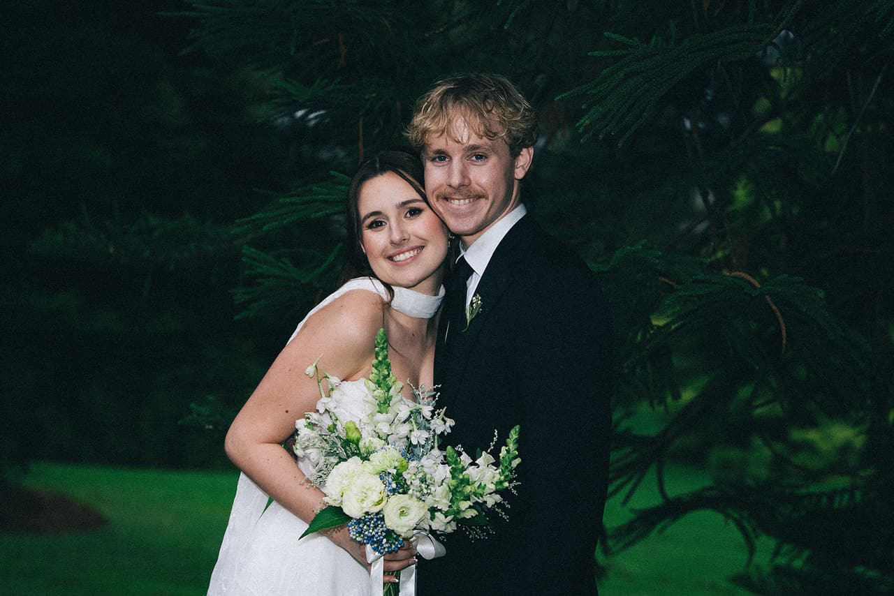 Charlotte and Eli cheek to cheek under the pine branches, her bouquet of white and blue flowers in hand