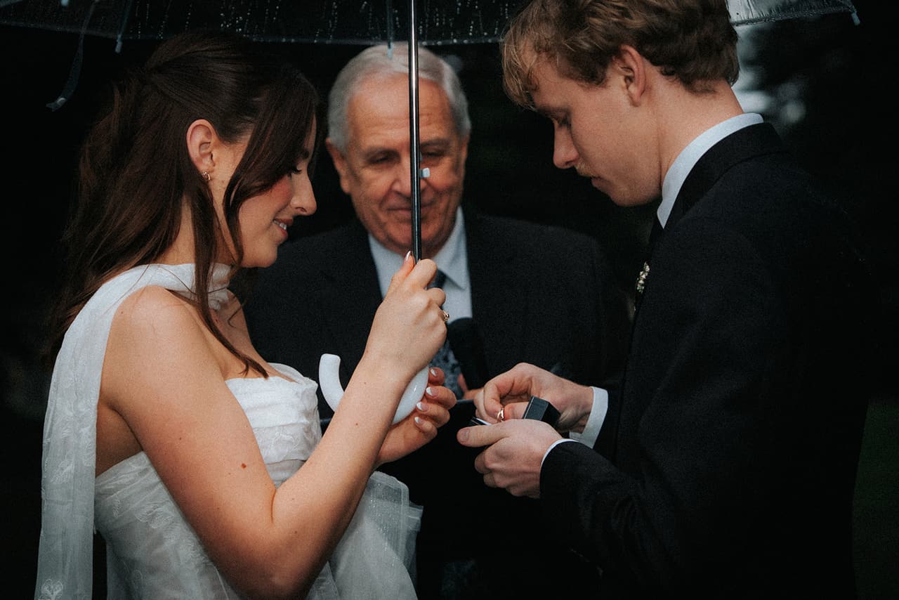 Charlotte smiling as Eli takes a ring from its box, the celebrant holding the umbrella over them