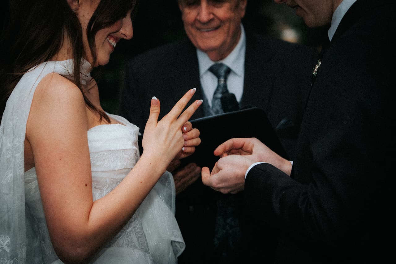 Charlotte smiling down at her hand during the ring exchange, Eli and the smiling celebrant beside her