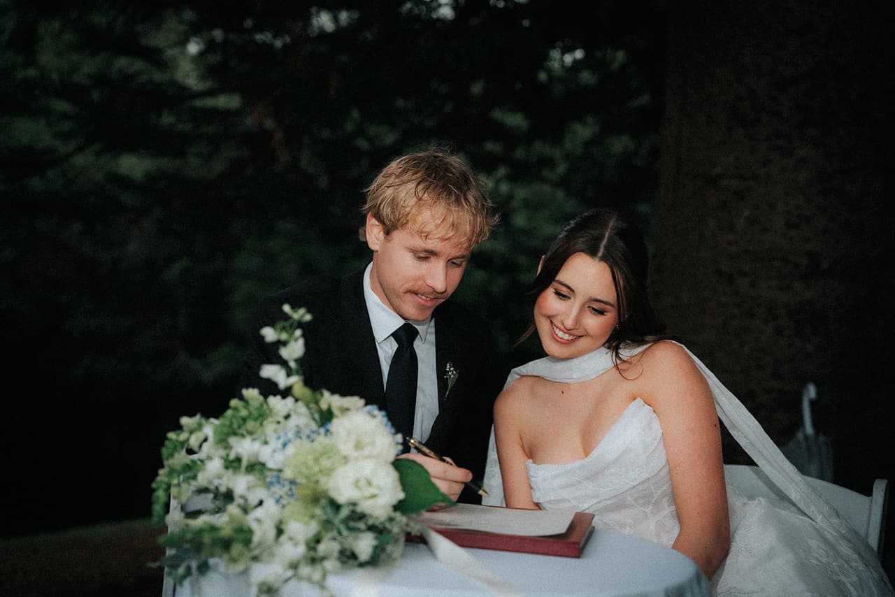 Eli signing the register at a small round table beside Charlotte, a bouquet of white flowers in front of them