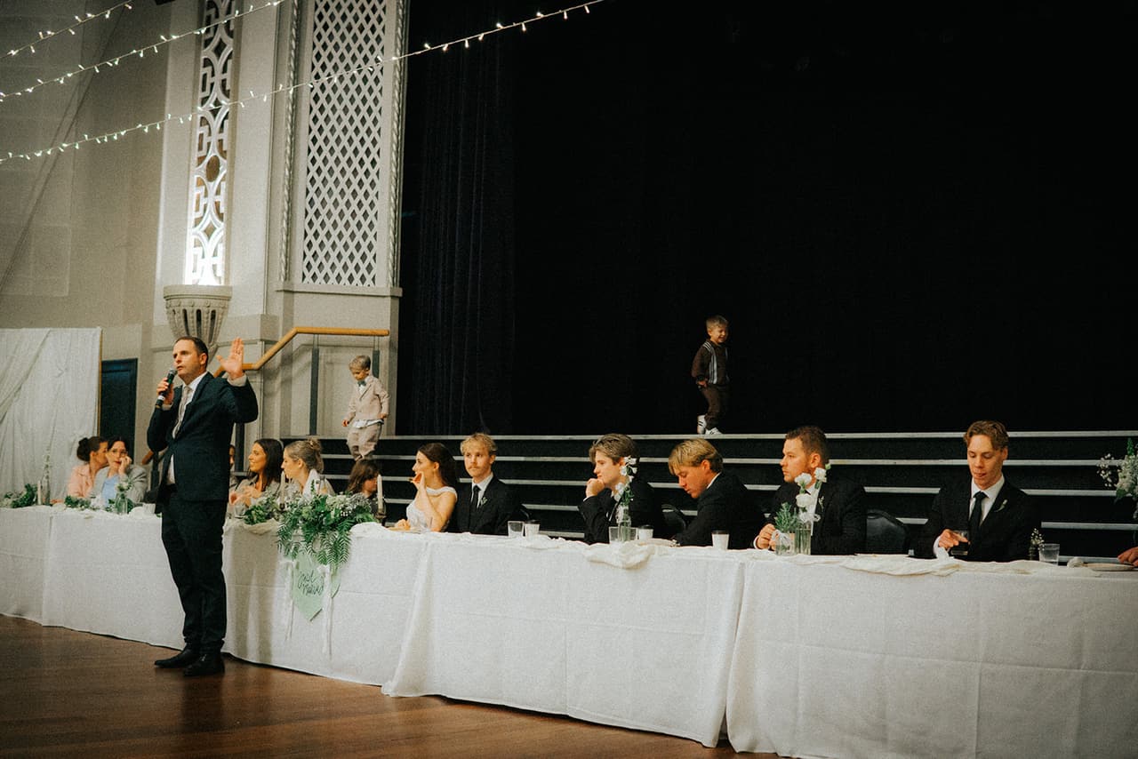 A man giving a speech into a microphone beside the long bridal table at Woodville Town Hall, the stage behind