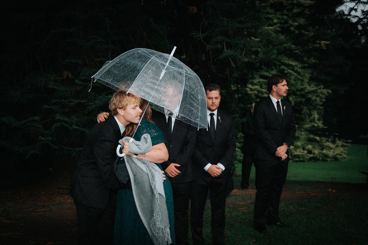 Eli hugging a woman in a green lace dress under a clear umbrella as the groomsmen wait behind him