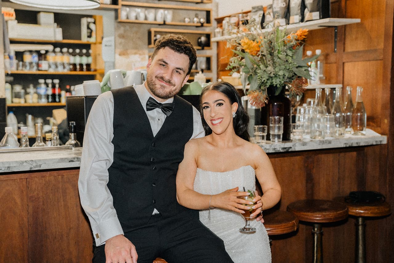 George and Tia perched on stools at the bar, shelves of bottles and flowers behind them