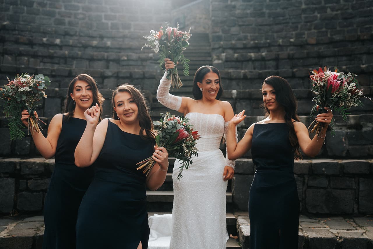 Tia and her three bridesmaids in navy raising their bouquets and cheering in the amphitheatre