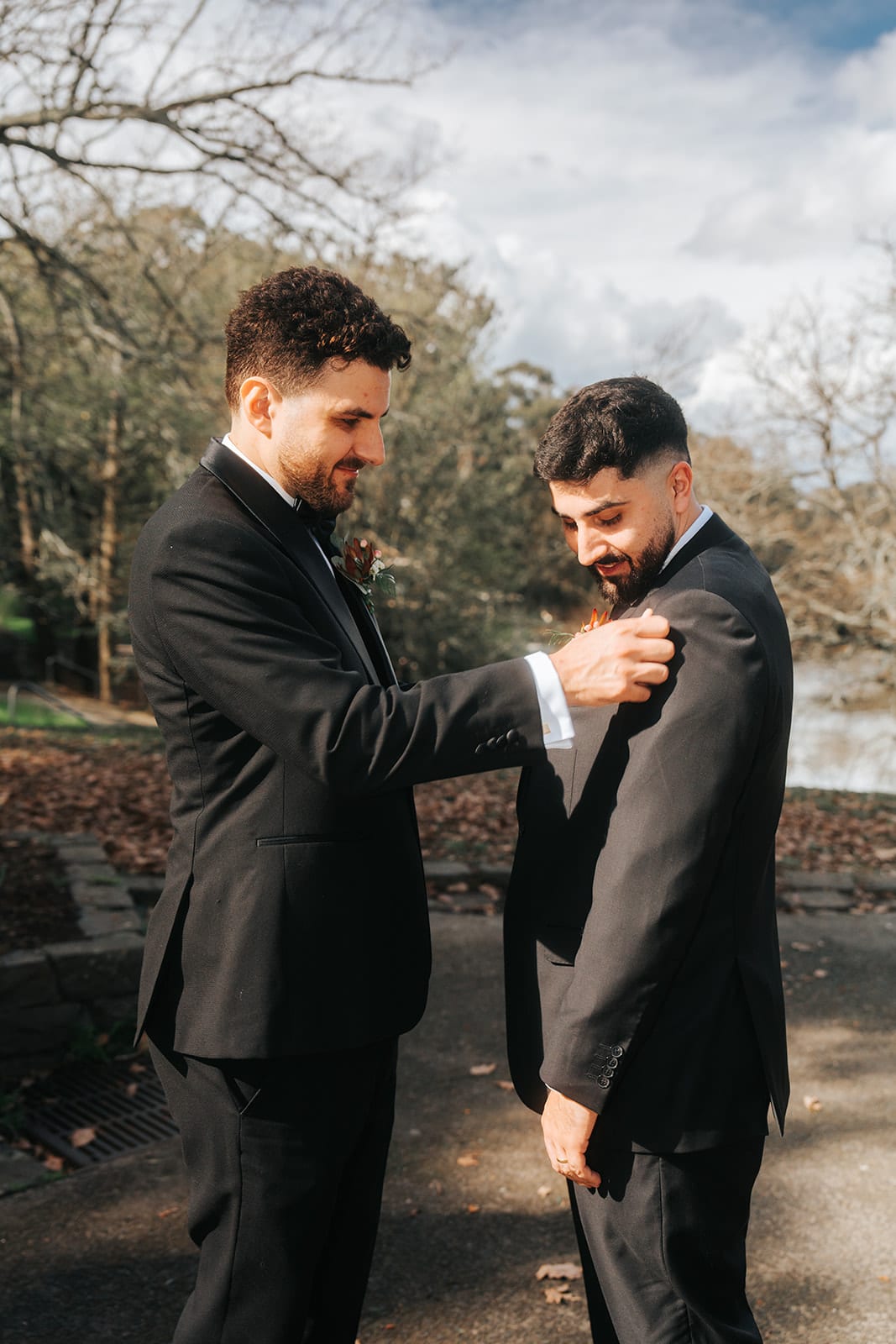 George pinning a buttonhole on one of his groomsmen under bare trees in the park