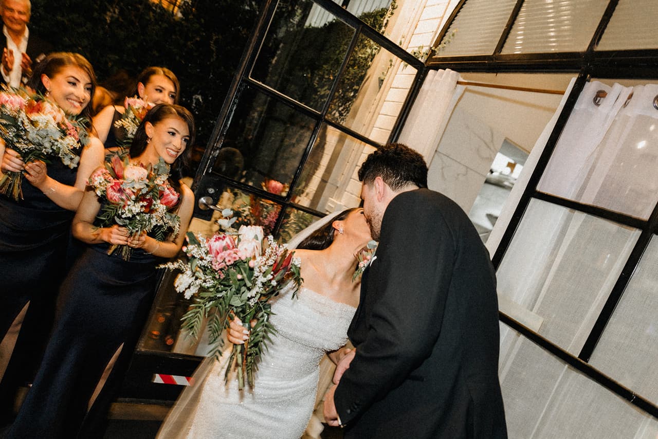 George kissing Tia at the black steel doors as her bridesmaids cheer with their bouquets