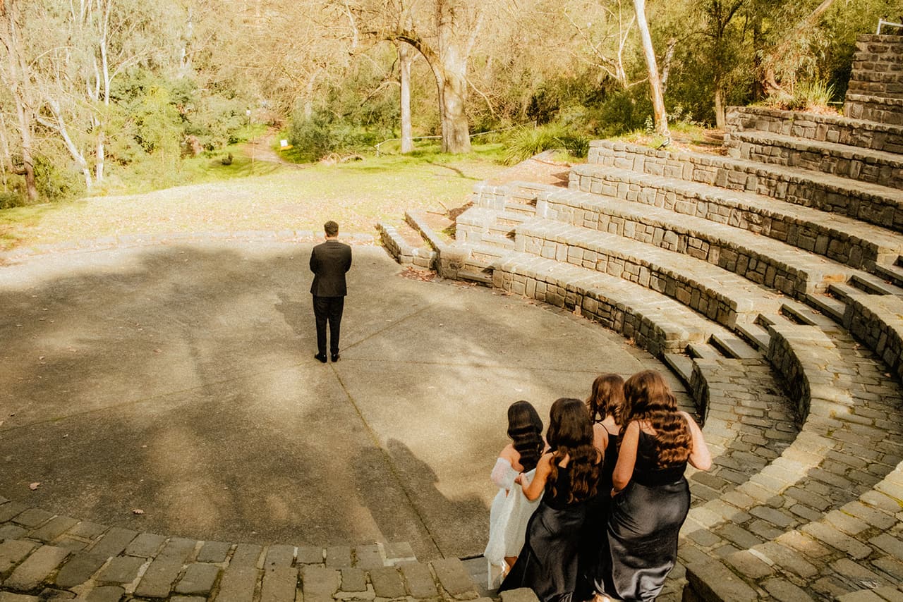 George waiting with his back turned in the middle of the amphitheatre while Tia and her bridesmaids gather on the terraces behind him