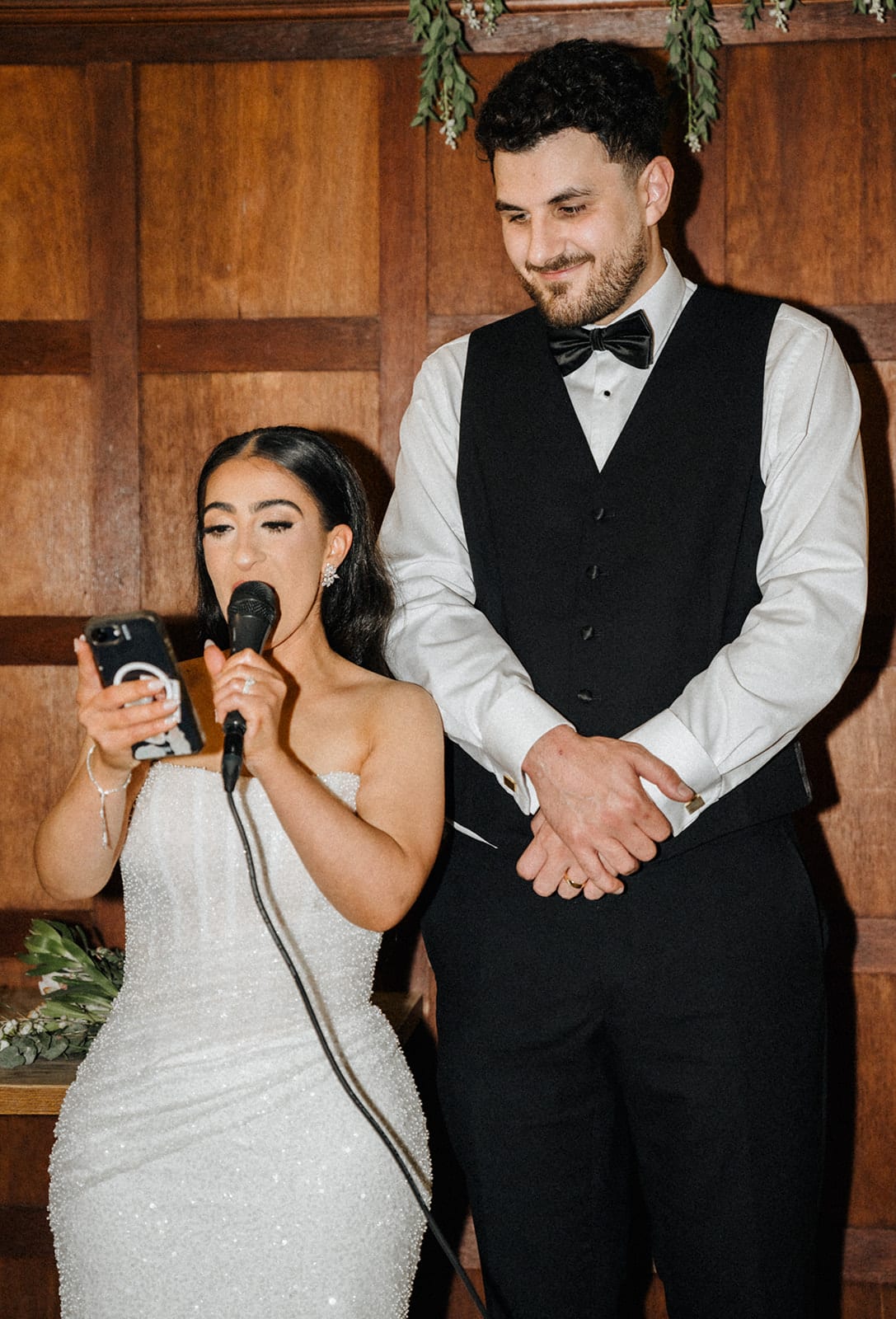 Tia giving her speech into a microphone, reading from her phone, as George listens beside her