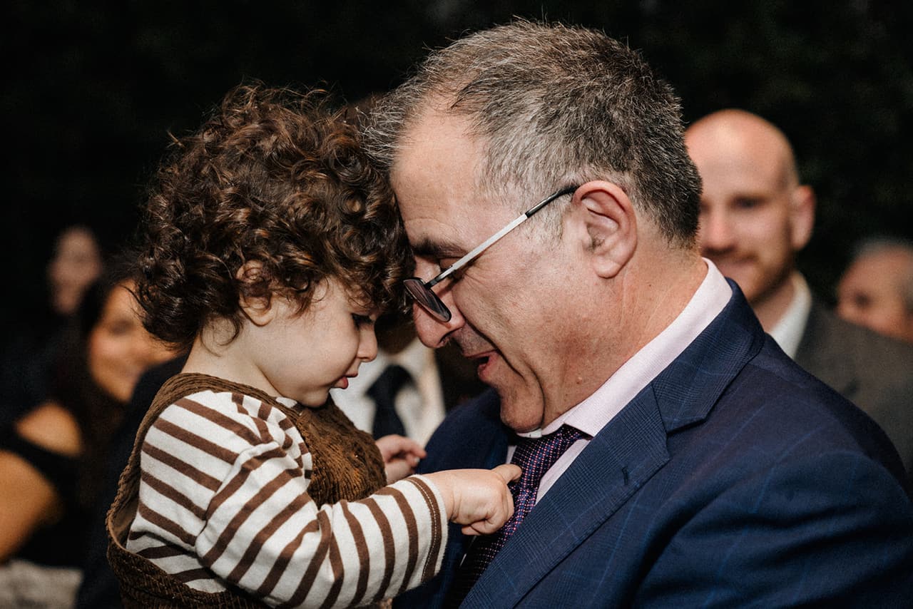 An older man in a blue suit touching foreheads with a curly-haired toddler as guests arrive