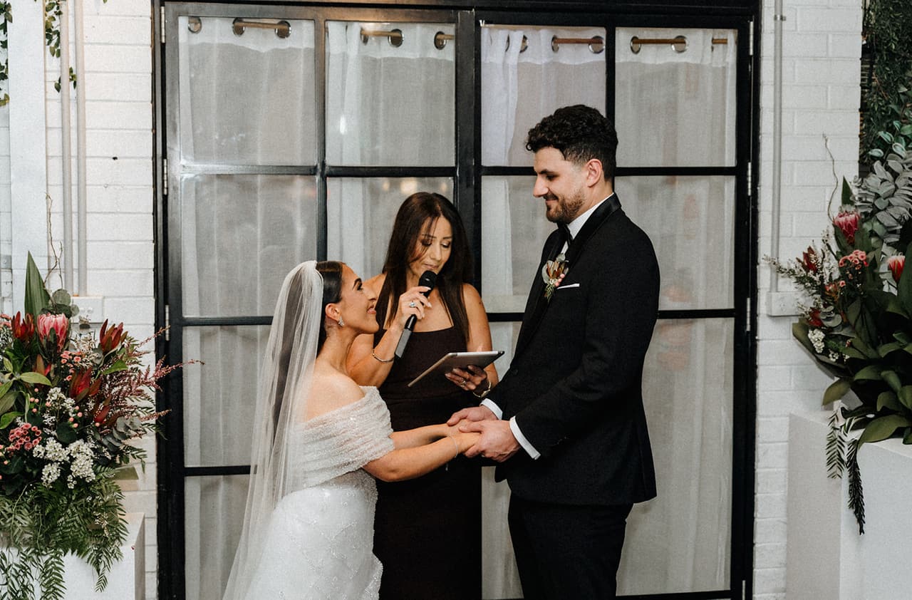 George and Tia saying their vows in front of a tall arched black window at Little Henri