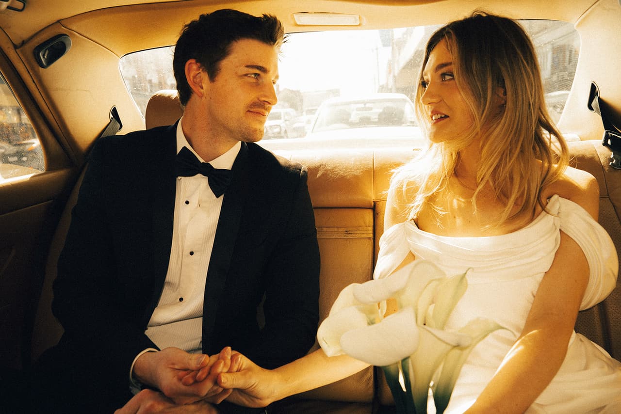 Hannah and Josh holding hands on the tan leather back seat of the Mercedes, calla lilies on her lap