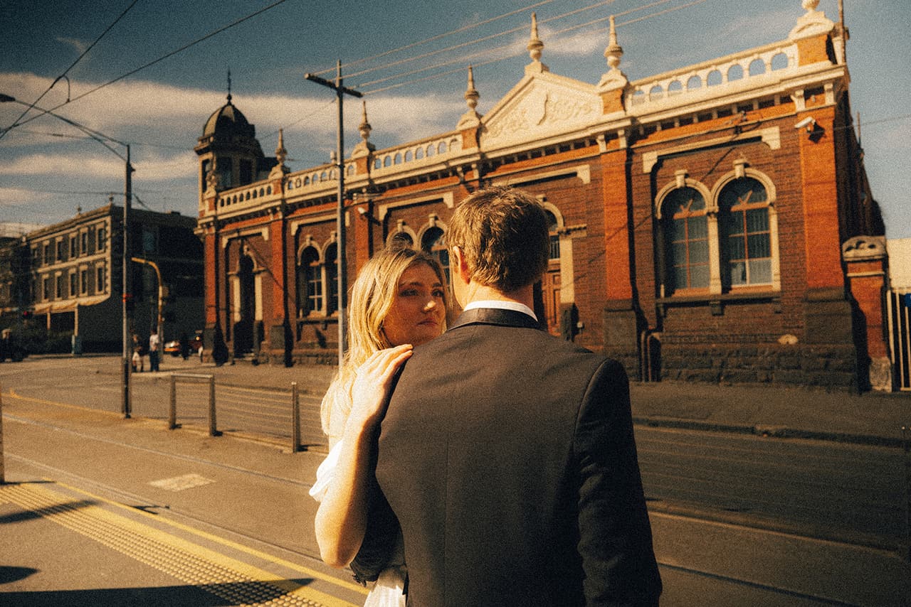 Hannah looking back over Josh’s shoulder across the tram tracks, an ornate red brick building with a turret behind them