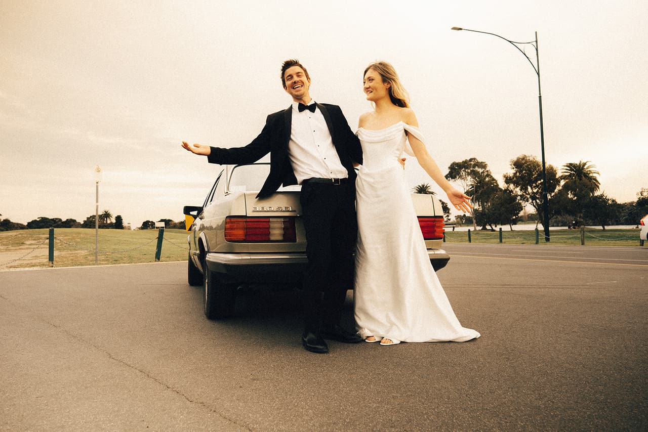 Josh throwing an arm out and Hannah laughing as they lean on the back of the Mercedes