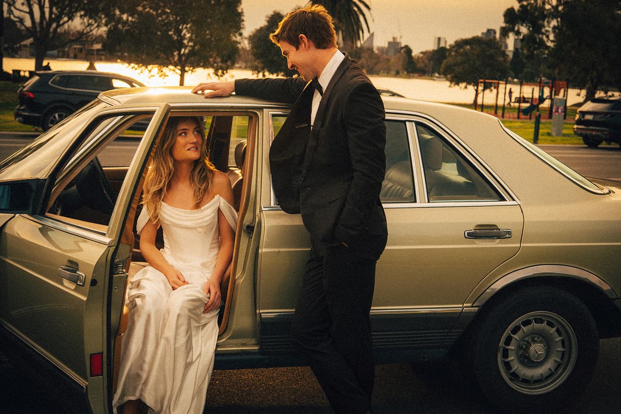Hannah sitting sideways in the open door of the Mercedes while Josh leans on the roof beside her