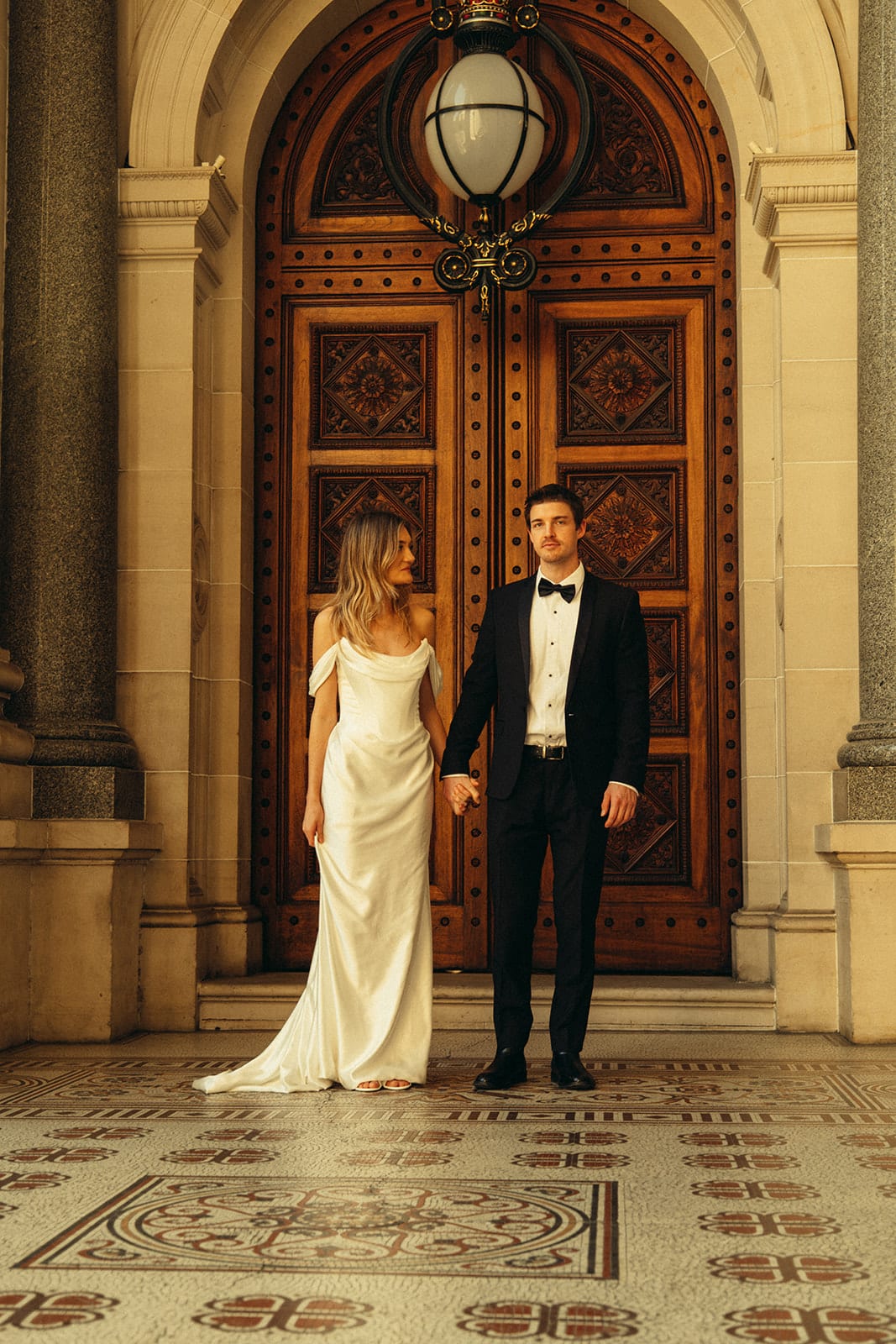 Hannah and Josh standing on the patterned tiles in front of the carved timber doors of Parliament House