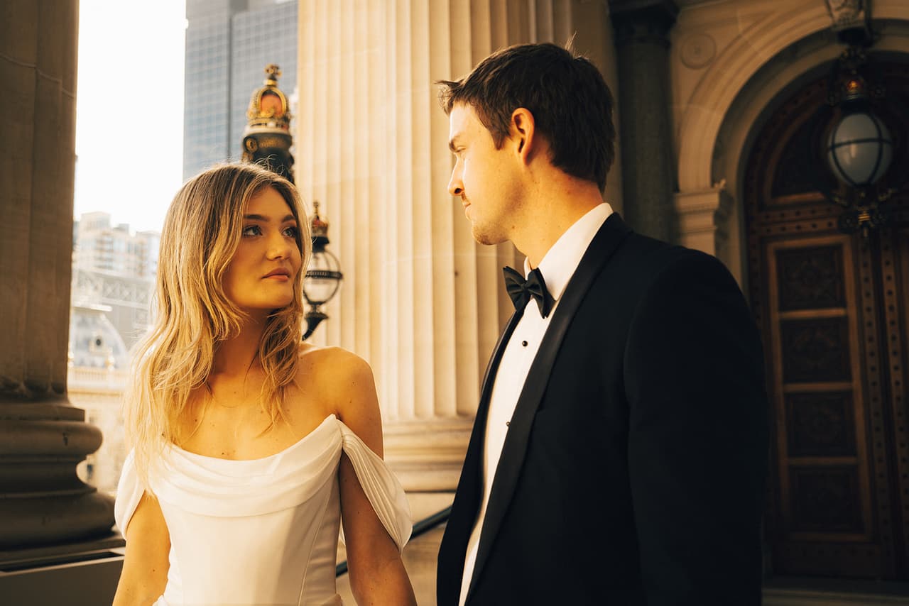 Hannah looking up at Josh between the columns of Parliament House, city towers beyond