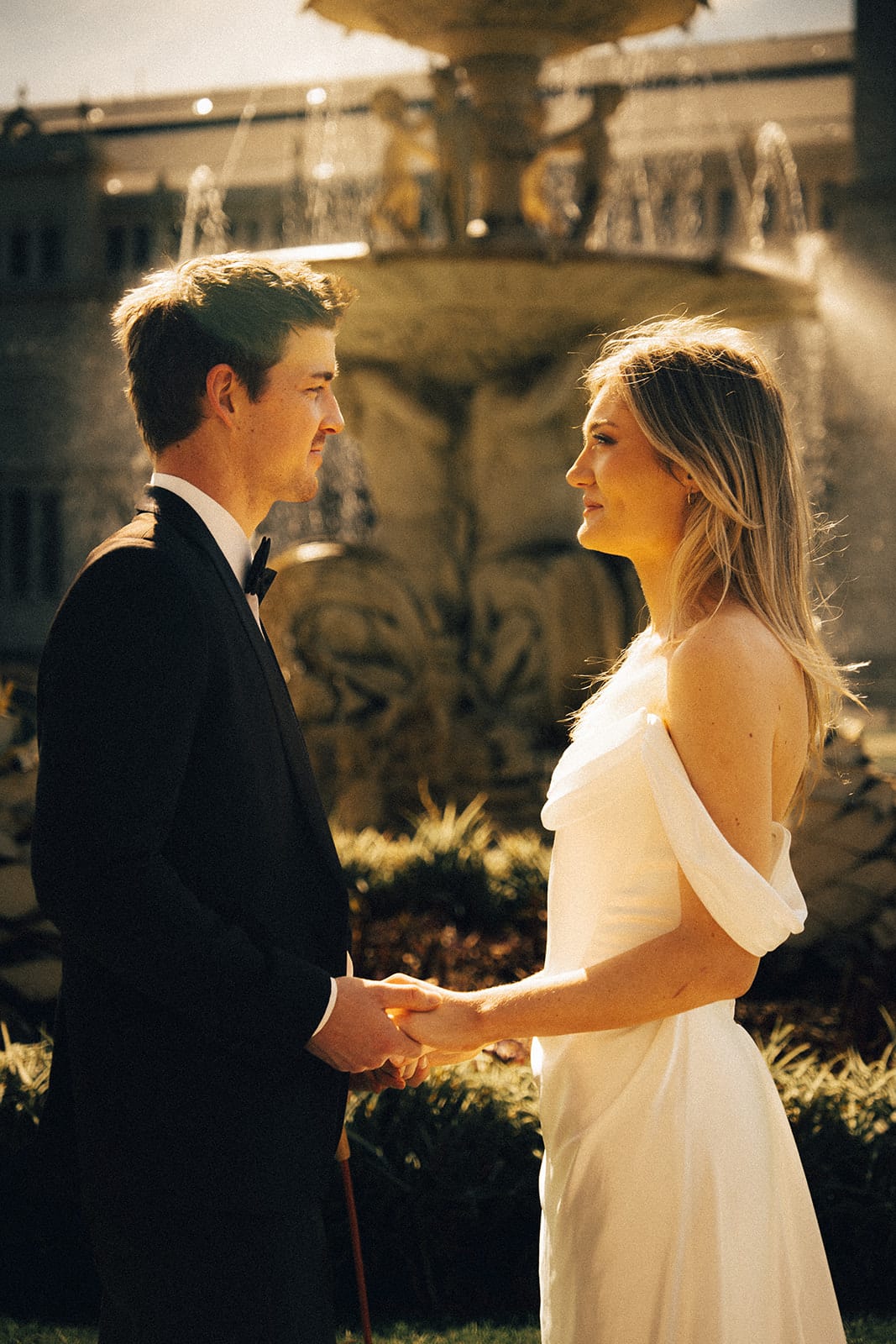 Hannah and Josh holding hands in front of the fountain, its spray lit by the sun behind them