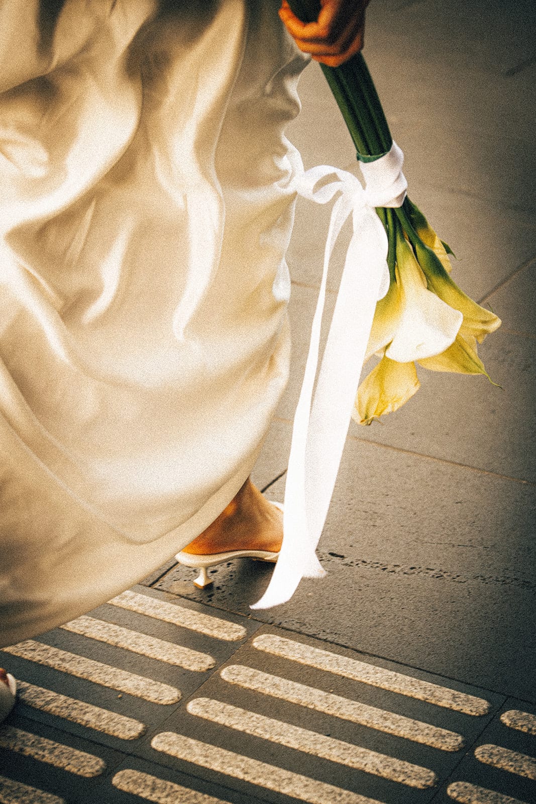 Close up of Hannah’s satin hem, white heels and calla lilies above the tactile paving of a crossing