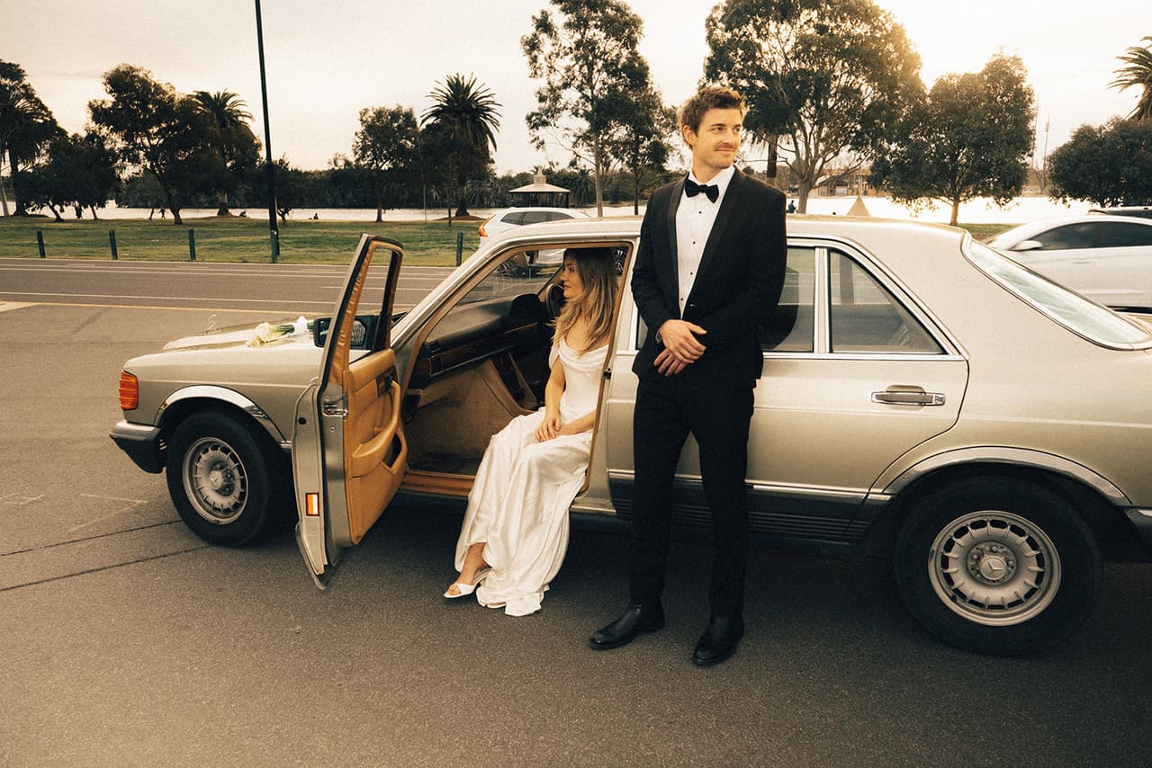 Hannah sitting in the open car door with Josh standing beside her, palms and parkland around the Mercedes