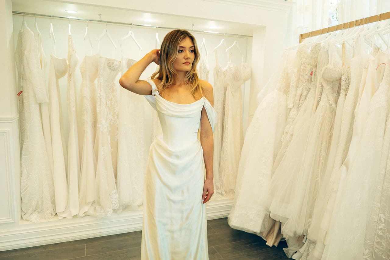 Hannah in her off-the-shoulder satin gown with a hand in her hair among racks of wedding dresses in a bridal shop
