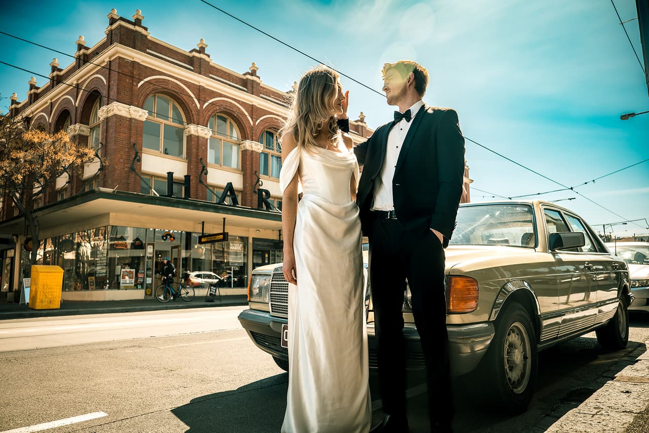 Hannah and Josh beside a vintage silver Mercedes in front of a red brick Victorian building under a blue sky