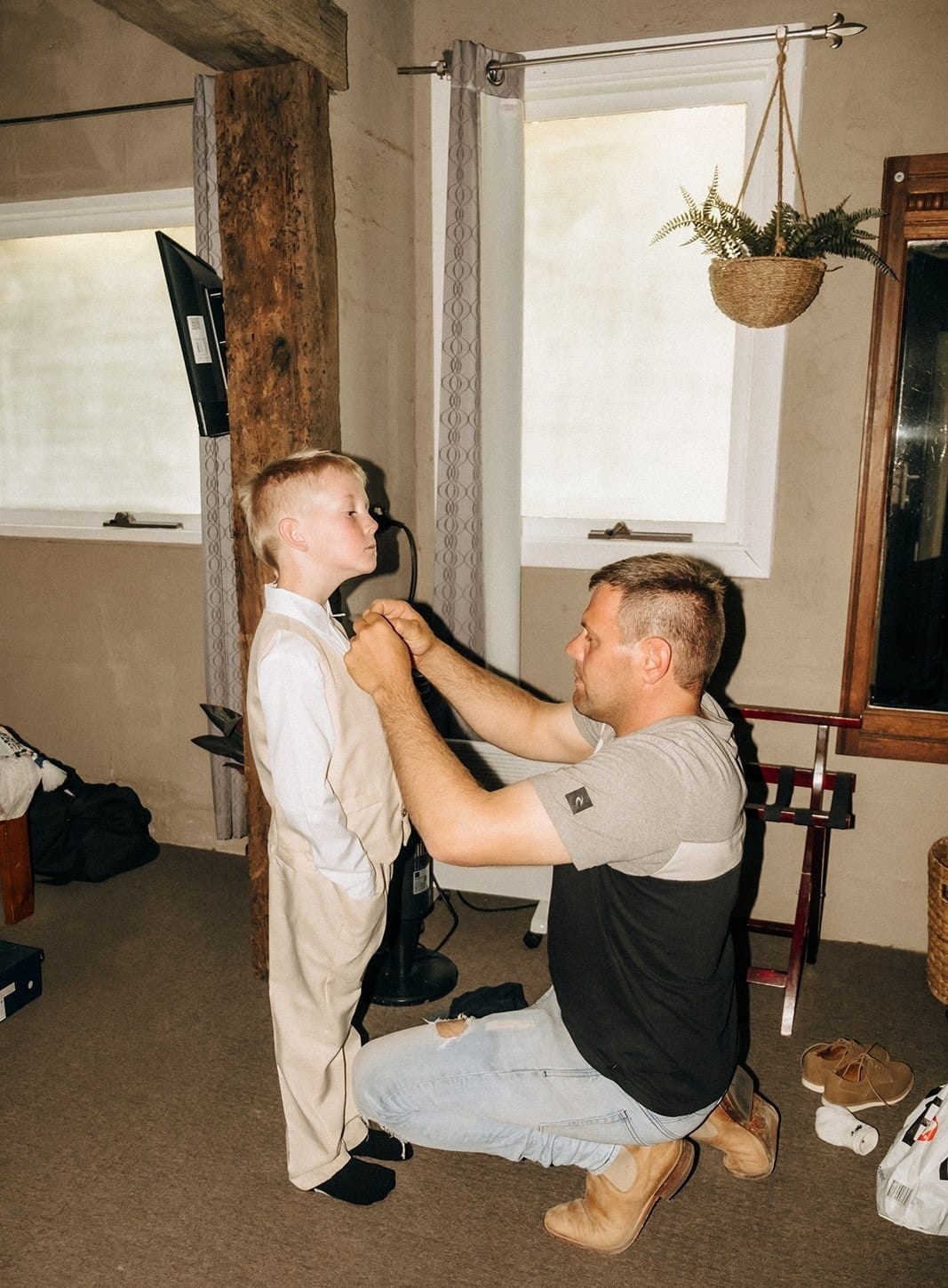 A man kneeling to help a page boy in a cream suit get dressed in the groom’s room