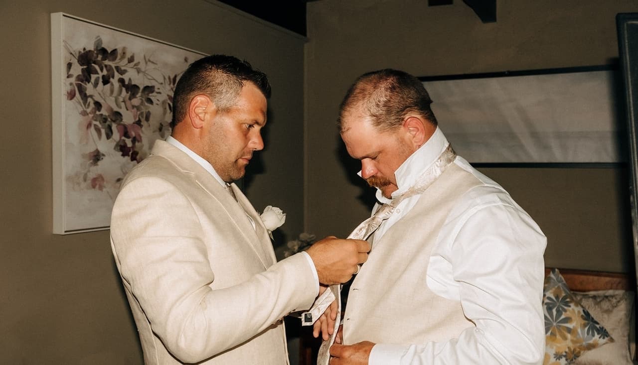 A white rose buttonhole being pinned on a cream jacket in the groom’s room