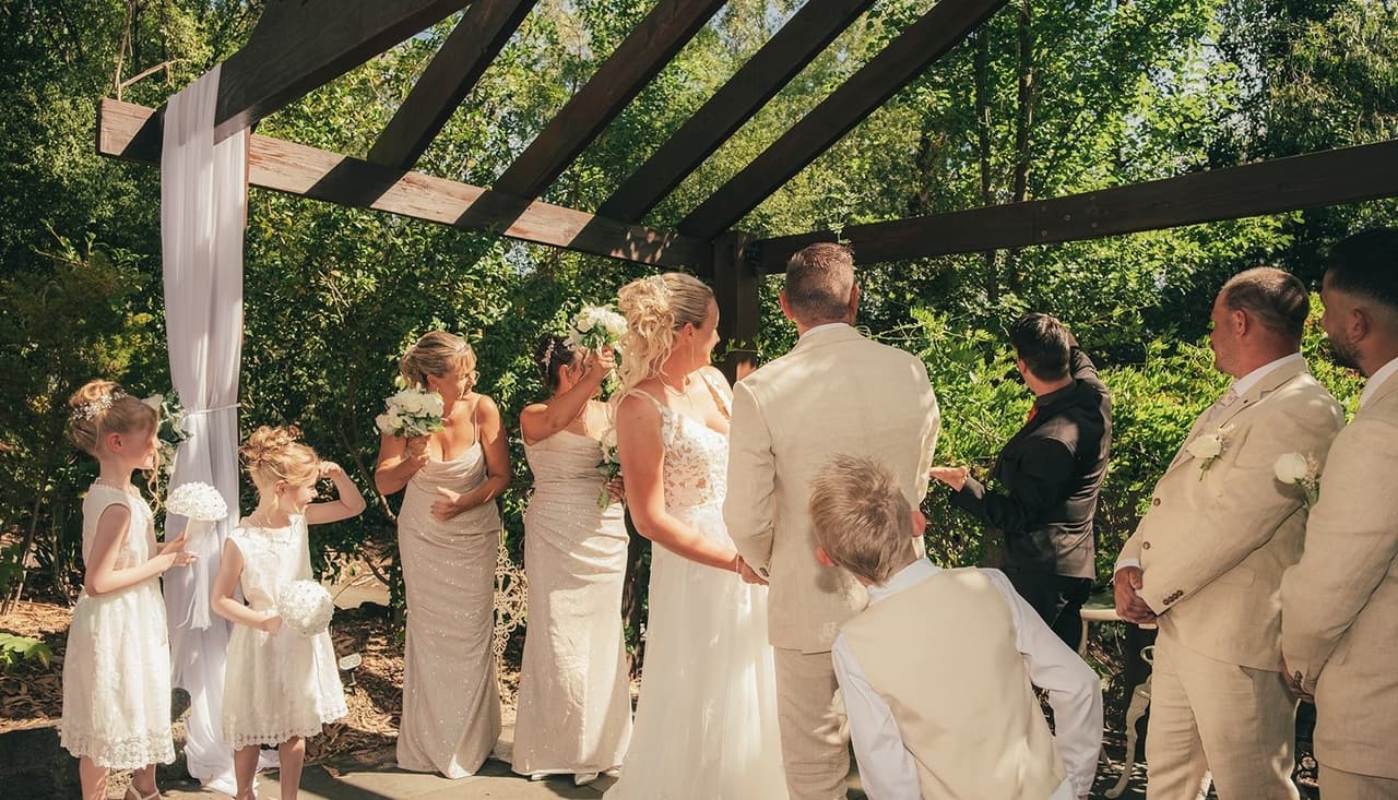 Jenna and Nick holding hands under the timber pergola with their bridesmaids, flower girls and groomsmen