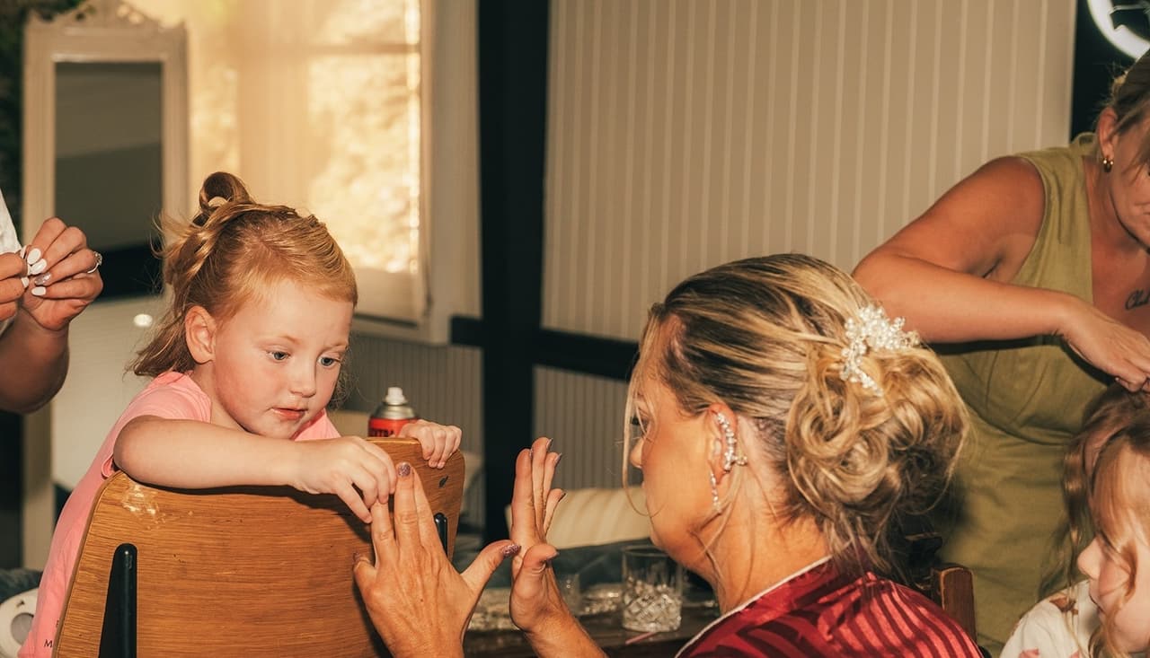 A little girl in a pink dress laughing with Jenna at the dressing table while her makeup is done
